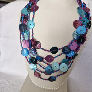 May include: Multi-strand necklace featuring blue, pink and purple round shell beads. The necklace is layered and has a variety of bead sizes.