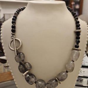 May include: A necklace with a toggle clasp, displayed on a white mannequin. The necklace features a strand of small, black beads and larger, grey, oval-shaped stones. Silver-toned metal accents and a large toggle clasp complete the design.