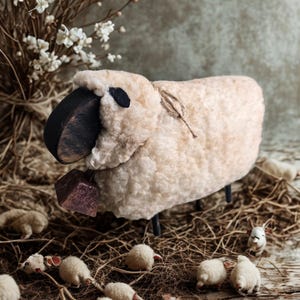 May include: A handcrafted sheep figurine with a fluffy, cream-colored body, black face, and small black legs. A small, purple bell hangs from its neck. Miniature sheep figurines are scattered around the base, creating a pastoral scene.