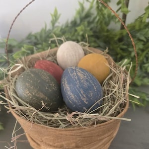 May include: A rustic basket filled with decorative Easter eggs. The eggs are painted in various colors, including green, blue, red, yellow, and white. The basket is lined with straw and has a wire handle. Greenery is visible in the background.
