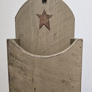 May include: A rustic, wooden wall pocket with a distressed finish. The pocket has a curved top and a cutout star. The color is a muted, neutral tone, with visible wood grain and a metal star.