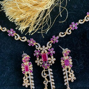 May include: A gold necklace and earrings set with a floral design and pink and white stones. The necklace has a tassel.