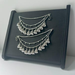 May include: A pair of silver ear chains with white pearl accents and dangling bells. The chains are layered and have a hook closure.