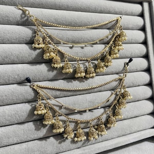 May include: Gold-toned ear chains with multiple strands. Each strand features small, bell-shaped charms with faux pearls. The chains are adorned with tiny, sparkling accents. The design is symmetrical, with a hook at the top and a clip at the bottom.