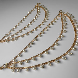 May include: Two sets of gold-toned, layered ear chains with dangling faux pearls. Each chain is composed of multiple strands, with small pearls evenly spaced along the lengths. The chains are connected at the top and bottom with a hook closure.