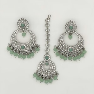 May include: A silver-coloured jewellery set comprising a pair of large, circular earrings and a headpiece. Each piece is decorated with small, clear crystals and light green beads. The earrings and headpiece have a floral design.