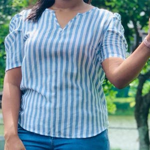 Könnte beinhalten: Ein blau-weiß gestreiftes Oberteil mit kurzen, gepufften Ärmeln und einem V-Ausschnitt. Das Oberteil wird mit einer blauen Jeans kombiniert. Das Bild wird im Freien mit einem verschwommenen grünen Hintergrund aufgenommen.