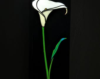 Puede incluir: Una pieza de arte mural en vitral que presenta un lirio de agua blanco con un espádice amarillo y un tallo y hoja de color verde vibrante. El diseño floral está colocado sobre un fondo rectangular negro sólido, creando un contraste visual.