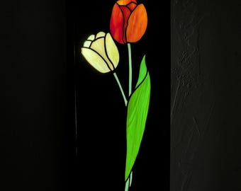 Puede incluir: Un panel rectangular de vidrieras que muestra dos tulipanes sobre un fondo negro sólido. Un tulipán es de un rojo anaranjado vibrante, mientras que el otro es de un amarillo crema pálido. Ambas flores tienen tallos verde brillante y hojas largas.