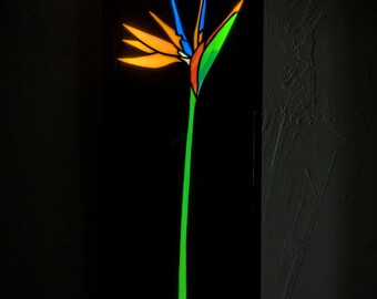 Puede incluir: Un panel de pared rectangular negro con un diseño de vidrieras retroiluminado de una flor ave del paraíso. La flor muestra segmentos vibrantes en naranja, azul y verde, mientras que el tallo largo está iluminado en un verde neón brillante.