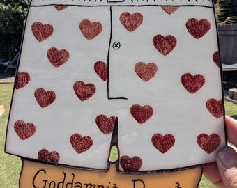Puede incluir: Una pieza de vitral blanco con forma de calzoncillos tipo bóxer con un patrón repetitivo de corazones rojos texturizados y un contorno negro. En la parte inferior, una cinta amarilla muestra el texto negro "Goddamnit Donut".