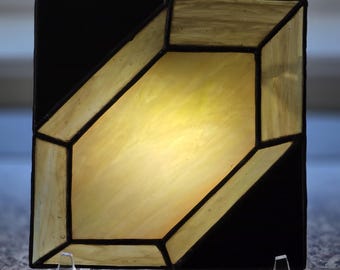 Puede incluir: Un panel de vidriera cuadrado que presenta una forma de cristal geométrica central en vidrio amarillo pálido translúcido. Las esquinas circundantes están compuestas por piezas de vidrio negro opaco, unidas por líneas de soldadura de metal oscuro.
