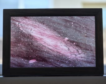 Puede incluir: Una obra de arte rectangular enmarcada que presenta un fondo oscuro y atmosférico con una franja diagonal de salpicaduras de pintura rosa suave y blanca que se asemeja a una nebulosa o galaxia. El marco es de material negro sólido.