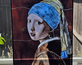 Puede incluir: Una pieza de arte en vidrio rectangular que presenta una representación al estilo de vidriera de una persona con un pañuelo azul brillante y un paño amarillo. La figura tiene cabello oscuro, labios rojos y un pendiente pequeño sobre fondo rojo.