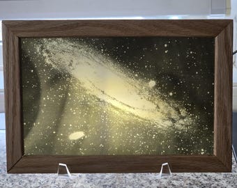 Puede incluir: Una obra de arte rectangular enmarcada que presenta una representación pintada con aerosol de una galaxia espiral sobre un fondo oscuro y moteado. La obra está encerrada en un marco de madera de tono medio y se exhibe en un soporte acrílico transparente.