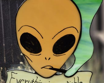 Puede incluir: Un atrapasoles de vidriera que muestra una cabeza alienígena amarilla con grandes ojos negros, fumando un cigarrillo negro con una estela de humo verde. Debajo de la cabeza hay una pancarta color crema con el texto negro Everything is negotiable.