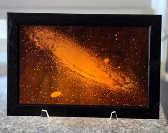Puede incluir: Una obra de arte rectangular enmarcada que presenta un diseño de galaxia espiral de color naranja vibrante sobre un fondo oscuro. La pieza está encerrada en un marco negro sencillo y se exhibe sobre un soporte de acrílico transparente.