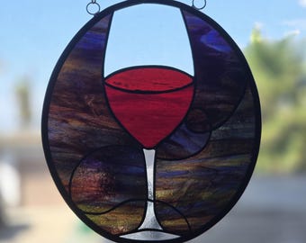 Puede incluir: Un atrapasoles circular de vidrieras con la silueta central de una copa de vino tinto. El fondo consta de segmentos de vidrio con remolinos de color púrpura oscuro, azul y ámbar, enmarcados en metal negro y suspendidos por una cadena metálica.