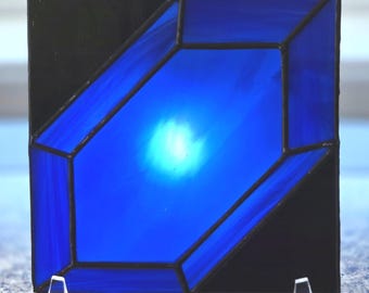 Puede incluir: Un panel de vidriera cuadrado que presenta una forma de cristal geométrica central en vidrio azul vibrante, rodeada por esquinas de vidrio negro sólido. Las piezas están unidas por líneas de soldadura de metal oscuro, creando un aspecto angular y moderno.