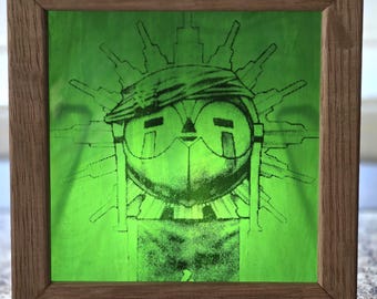 Puede incluir: Un marco de madera cuadrado que contiene un panel translúcido de color verde brillante. El panel presenta un estampado gráfico negro de una figura estilizada con un tocado y gafas redondas grandes, rodeada por un patrón radiante de líneas verticales.