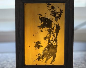 Puede incluir: Una caja de luz rectangular con un marco de madera oscura y un panel de color ámbar brillante. El panel muestra la silueta negra de alto contraste de una figura de perfil, formada por salpicaduras abstractas y texturizadas tipo tinta.