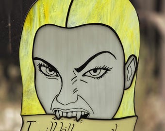 Puede incluir: Un atrapasoles de vidriera que presenta un rostro estilizado con cabello amarillo brillante, piel pálida y colmillos afilados. La figura sostiene una pancarta de color amarillo claro en la boca con el texto negro "I will kill your mother" en cursiva.