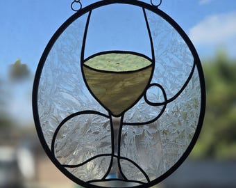 Puede incluir: Un atrapasoles circular de vidrieras con una copa de vino amarilla central sobre un fondo texturizado y transparente con patrón de escarcha. La pieza está enmarcada en un borde de metal negro y cuelga de una cadena de tono plateado.