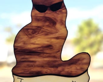 Puede incluir: Un atrapasoles de vidriera con forma de gato sentado, hecho de vidrio veteado marrón, gafas de sol de vidrio negro y una banda color crema en la base con el texto negro "I mean, really". La pieza cuelga de una cadena de metal plateado.