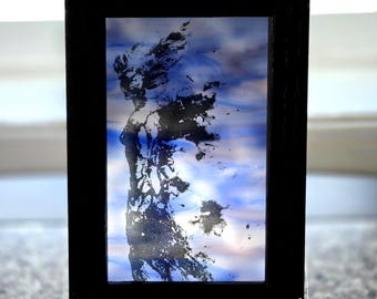 Puede incluir: Un marco rectangular negro que contiene un panel translúcido con un fondo jaspeado en azul y blanco. Una silueta oscura y texturizada de una figura con cabello o alas que fluyen está centrada en el panel, creando un efecto visual contrastado.