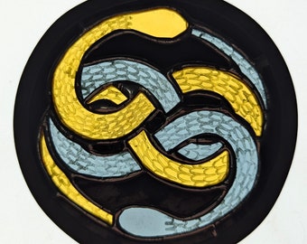 Peut inclure: Un motif de vitrail circulaire représentant deux serpents entrelacés, l'un jaune et l'autre bleu, sur fond noir.