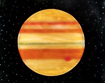 Puede incluir: Una pieza de arte circular en vitral que representa al planeta Júpiter con bandas horizontales de color amarillo, naranja y rojo oscuro, incluyendo una pequeña mancha roja. El planeta se sitúa sobre un fondo negro con puntos blancos pintados.