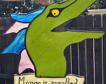 Puede incluir: Un atrapasoles de vidrieras con forma de cabeza de criatura verde con un ojo amarillo, aletas azules y rosas, y una pancarta color crema en la parte inferior con el texto negro 'Mongo is appalled' delineado por líneas de soldadura de metal oscuro.