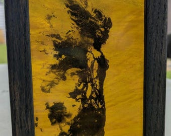 Peut inclure: Une œuvre d'art encadrée avec une silhouette noire sur un fond jaune. La silhouette représente une figure avec des cheveux flottants ou une coiffe. Le cadre marron foncé entoure l'œuvre, qui a une finition texturée.