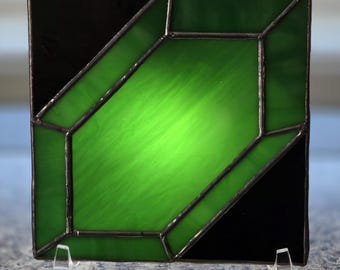 Puede incluir: Un panel de vidriera cuadrado que presenta una forma geométrica central de color verde esmeralda con apariencia facetada, rodeada por esquinas de vidrio negro sólido. Las piezas de vidrio están unidas por líneas de soldadura de metal oscuro.