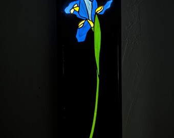 Puede incluir: Un aplique de pared rectangular negro con una flor de iris de vidriera retroiluminada. La flor tiene pétalos de color azul vibrante con detalles en amarillo brillante y un tallo verde largo y delgado, iluminado sobre un fondo negro.