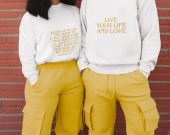 May include: Two people wearing matching white sweatshirts and yellow cargo pants. One sweatshirt has the text "LIGHT UP MY LIFE AND SHOW ME LOVE...WE NEED IS LOYALTY" in gold. The other sweatshirt says "LIVE YOUR LIFE AND LOVE" in gold.