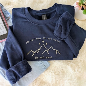 May include: Navy blue sweatshirt with the text "Do not fear, Do not falter, Do not yield" above a mountain graphic. The sweatshirt is folded on a fluffy white surface, with a pair of light blue jeans and a small potted plant visible.