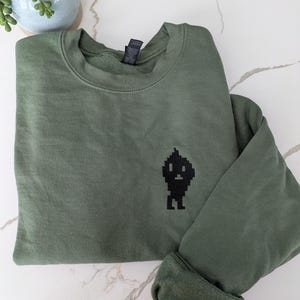 May include: An olive green sweatshirt with a black pixelated graphic on the front. The long-sleeved shirt is folded, showing the rolled-up cuff. The sweatshirt is made of a soft material.