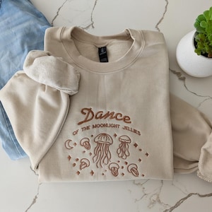 May include: A beige crewneck sweatshirt featuring brown embroidery that reads "Dance OF THE MOONLIGHT JELLIES" above a design of various jellyfish, stars, and a crescent moon. The sweatshirt is displayed alongside light blue denim jeans.