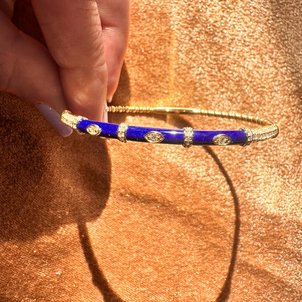 Pode incluir: Pulseira de bracelete dourada com uma seção central em esmalte azul e detalhes brilhantes de cristal. A pulseira é segurada por uma mão, mostrando seu design intrincado e apelo elegante. Uma peça de joalheria elegante.