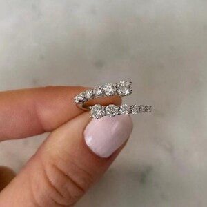 May include: A bypass ring crafted from polished silver-toned metal, featuring two open-ended bands that overlap. Each band is set with a row of five round, clear, faceted stones that catch the light, creating a continuous sparkling effect.
