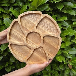 Op de afbeelding: Een ronde houten serveerschaal met meerdere compartimenten. Het lichte hout heeft een natuurlijk nerfpatroon. De compartimenten zijn in verschillende vormen en maten, ideaal voor het serveren van snacks of hapjes. De schaal wordt door twee handen vastgehouden tegen een achtergrond van groene bladeren.