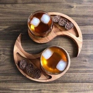 May include: Two glasses of amber liquid with ice cubes sit on wooden coasters, each placed on a matching wooden tray. The trays are crescent-shaped with a small indentation holding three chocolate candies. The background is a wooden surface.