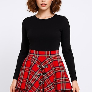Puede incluir: Una camiseta negra de manga larga combinada con una falda a cuadros roja. La falda presenta un dobladillo asimétrico único y hebillas decorativas. El patrón de cuadros incluye líneas rojas, amarillas y azul marino.