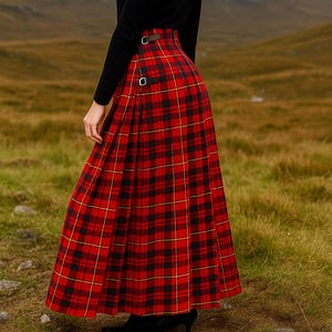 Op de afbeelding: Een lange, rood-zwarte geruite rok met een hoge taille en gespdetails. De rok wordt gecombineerd met een zwarte top met lange mouwen. De achtergrond toont een bergketen en een graslandschap.