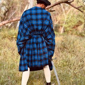 Può includere: Una persona indossa un kilt a quadri blu e neri e un mantello abbinato, un basco nero, calzini bianchi e stivali neri. Una spada d'argento è appoggiata a terra. La persona è in piedi in un campo erboso.