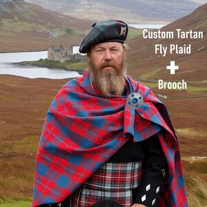 Puede incluir: Un hombre con un fly plaid tartán rojo y azul, boina y broche. La imagen incluye el texto "Custom Tartan Fly Plaid + Brooch". El fondo muestra un castillo y un lago.