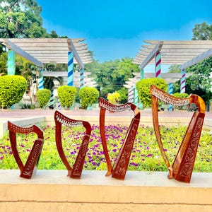 Può includere: Quattro arpe in legno di diverse dimensioni sono esposte all'aperto. Le arpe presentano intagli intricati ed elementi decorativi sulle loro tavole armoniche. Lo sfondo mostra un parco con vegetazione e cielo blu, suggerendo un tema musicale.