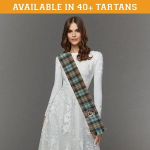 May include: A woman wearing a white dress with a long, plaid sash. The sash is green, brown, and yellow with a circular design in the center. The text "AVAILABLE IN 40+ TARTANS" is at the top of the image.