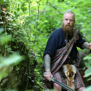 May include: A man with a long beard and a kilt is standing in a woodland. He is holding a sword in one hand and an axe in the other. He is wearing a black shirt and a kilt with a red and green tartan pattern. The kilt has a fox fur attached to it.